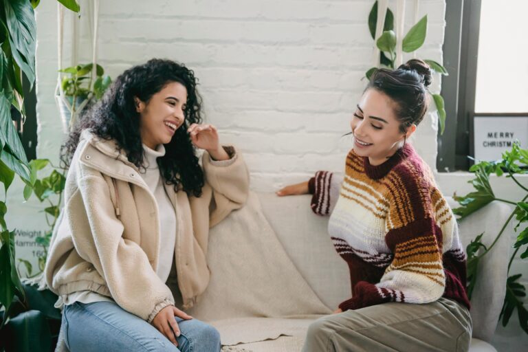 Two friends laughing on a couch, representing the surface harmony of people pleasing in friendships.