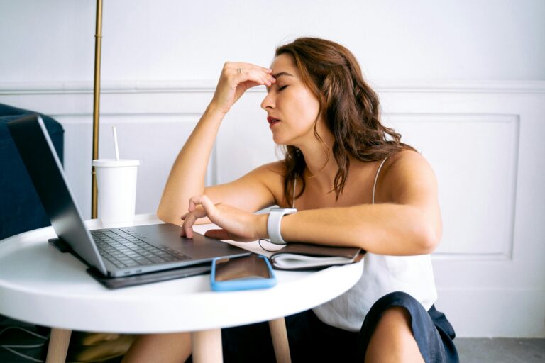 Woman at a table with a laptop and phone, hand pressed to her forehead and eyes closed — illustrating the nervous system and stress response activating in an ordinary moment.