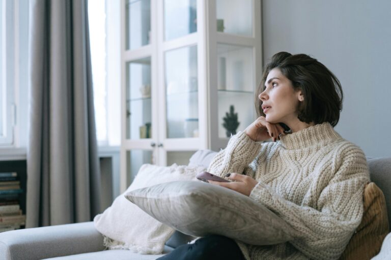 Woman sitting on a sofa holding a phone, looking away thoughtfully with her hand to her chin — illustrating the prefrontal cortex pausing to think before responding.