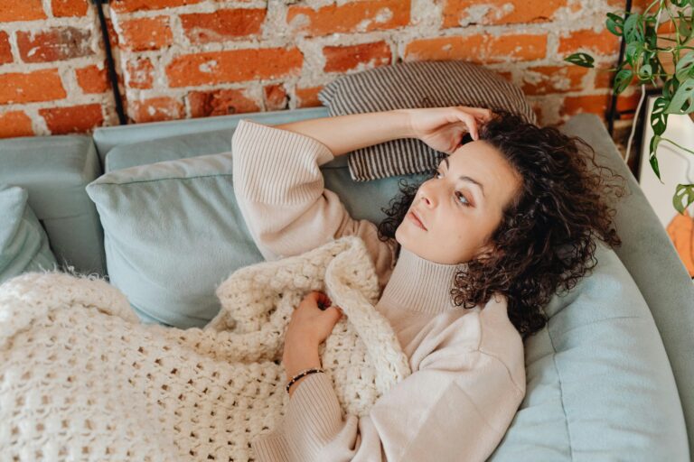 Burnout. Woman lying on a sofa wrapped in a blanket, eyes open and staring upward — illustrating the burnout experience of resting without recovering.