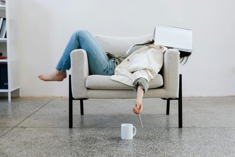 avoidance vs procrastination — Person slumped in a chair with a notebook covering their face and a pen dangling from their hand — illustrating the procrastination experience of having everything ready but not starting.