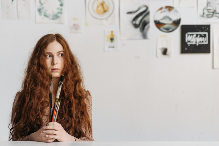 Young woman holding paintbrushes at a table with artwork pinned to the wall behind her, looking sideways with hesitation — illustrating the self-sabotage pattern of holding back when everything is in place to begin.