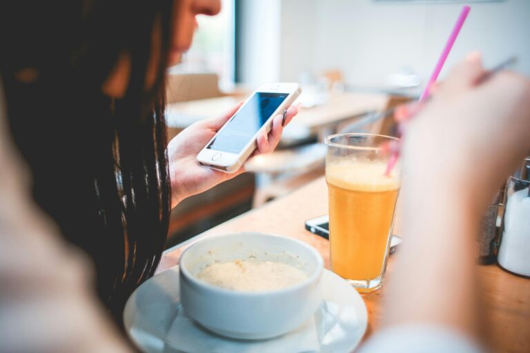 Woman checking her phone at a café with food and a drink in front of her — illustrating how dopamine-driven behavior pulls attention away from the present moment.
