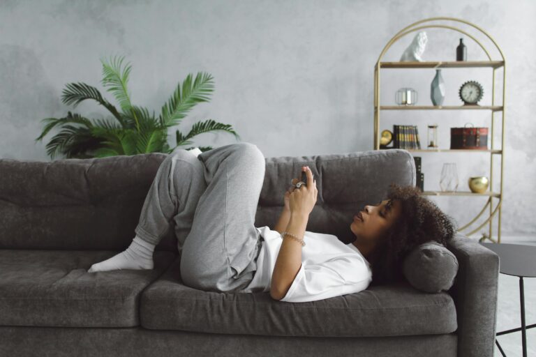 Woman lying on a sofa scrolling her phone with a clock visible on the shelf behind her — illustrating the avoidance pattern of choosing something easier while time passes.