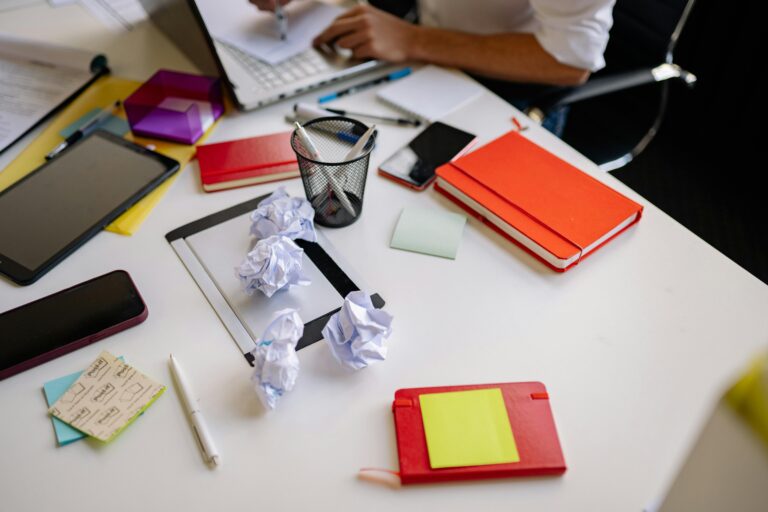 how to work with perfectionism - A cluttered desk with crumpled paper balls, notebooks, and sticky notes — illustrating the cycle of redoing and never feeling finished that perfectionism creates.