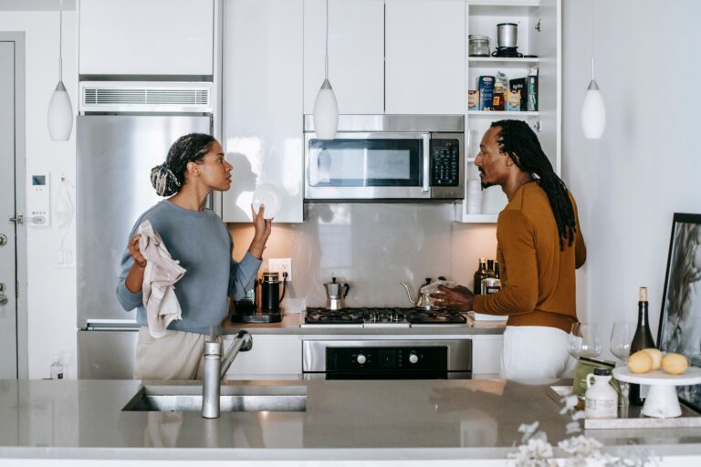 Couple facing each other in their kitchen, one holding up a plate mid-conversation, illustrating how a small comment about something like dishes can carry far more weight than the task itself.