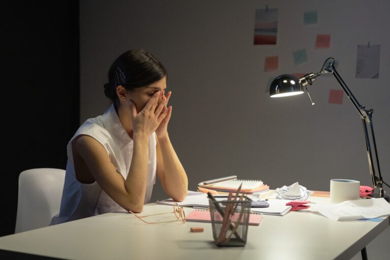 Woman sitting at a late-night desk with her face in her hands, surrounded by work, illustrating the exhaustion of working hard at something that never quite feels like enough.
