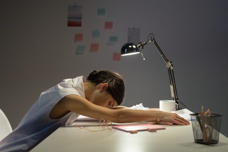 Woman with her head down on a desk surrounded by work materials, illustrating the exhaustion of repeated effort that leads to learned helplessness and stopping trying.