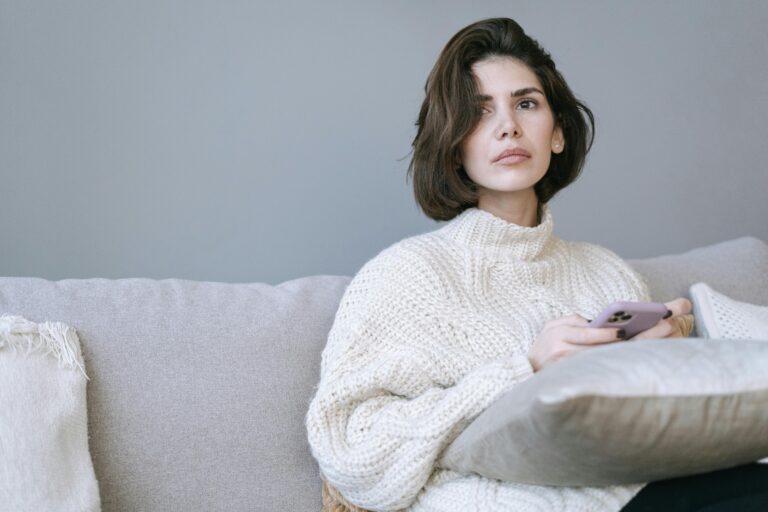Woman sitting on a sofa holding her phone with a blank, unfocused expression, illustrating the freeze response — going still and disconnected before any conscious decision is made.