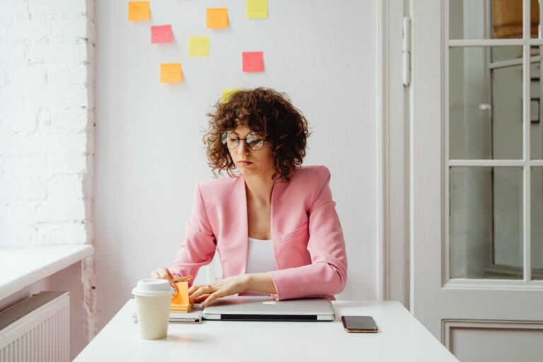 Woman sitting at a desk with colorful sticky notes on the wall behind her, illustrating the practice of stating your next action before doing it.