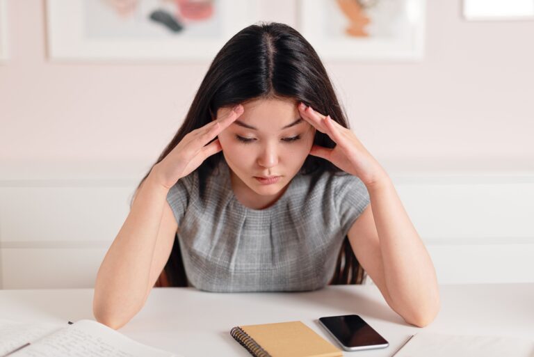 Woman pressing her fingers to her temples at a desk, illustrating the overlap between anxiety and overthinking — both the mental loop and the physical tension running at the same time.