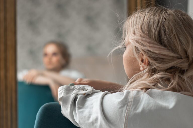 Woman looking at her own blurred reflection in a mirror, illustrating how avoidance blocks the experiences that shape your sense of self over time.