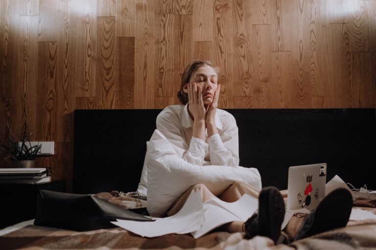 Person sitting on a bed with eyes closed and hands holding their face, surrounded by work materials, illustrating the exhaustion and mental fog caused by chronic stress on the nervous system.