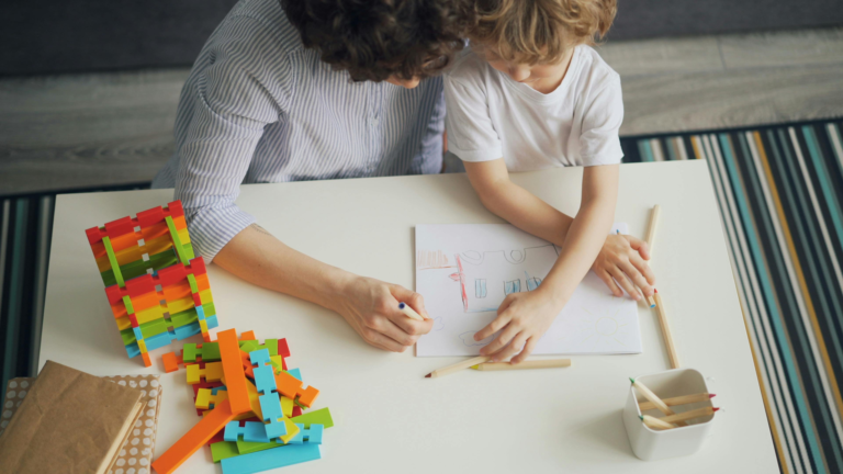 An adult and child sit side by side at a table, drawing together with colored pencils, with colorful building blocks nearby.