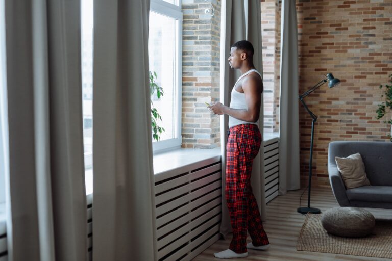Man standing at a window holding his phone with a distant expression, illustrating decision paralysis — the moment of being stuck between knowing a decision needs to be made and not being able to make it.