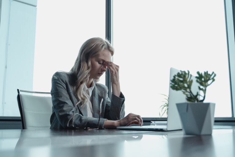Woman pinching the bridge of her nose at her desk with her eyes closed, illustrating the private moment of shame that often sits underneath perfectionism, invisible to anyone else in the room.