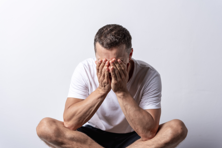 Man sitting on the floor with hands covering his face, illustrating what anxiety feels like in the body — the nervous system on alert when nothing around you explains why.