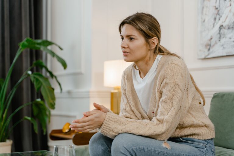 Woman sitting on the edge of a couch with hands clasped and gaze averted, illustrating the feeling of shame in a social setting — the moment you start watching yourself from outside and measuring every word before you say it.