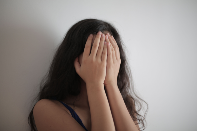 Woman pressing both hands over her face against a white wall, illustrating the state of being too overwhelmed for calming techniques to reach — when the system has gone past its own limit.