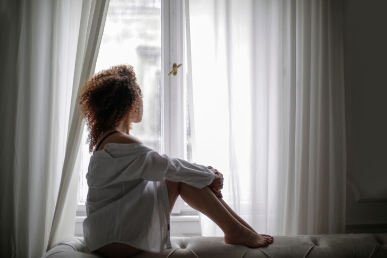 Woman sitting alone with her back to the camera, looking out a window, illustrating the quiet and constant internal weight of chronic shame — present even when nothing on the outside shows it.