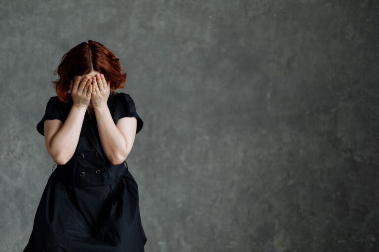 Woman pressing both hands over her face, head bowed, illustrating the physical sensation of a shame response — the body closing in before any thought has caught up.