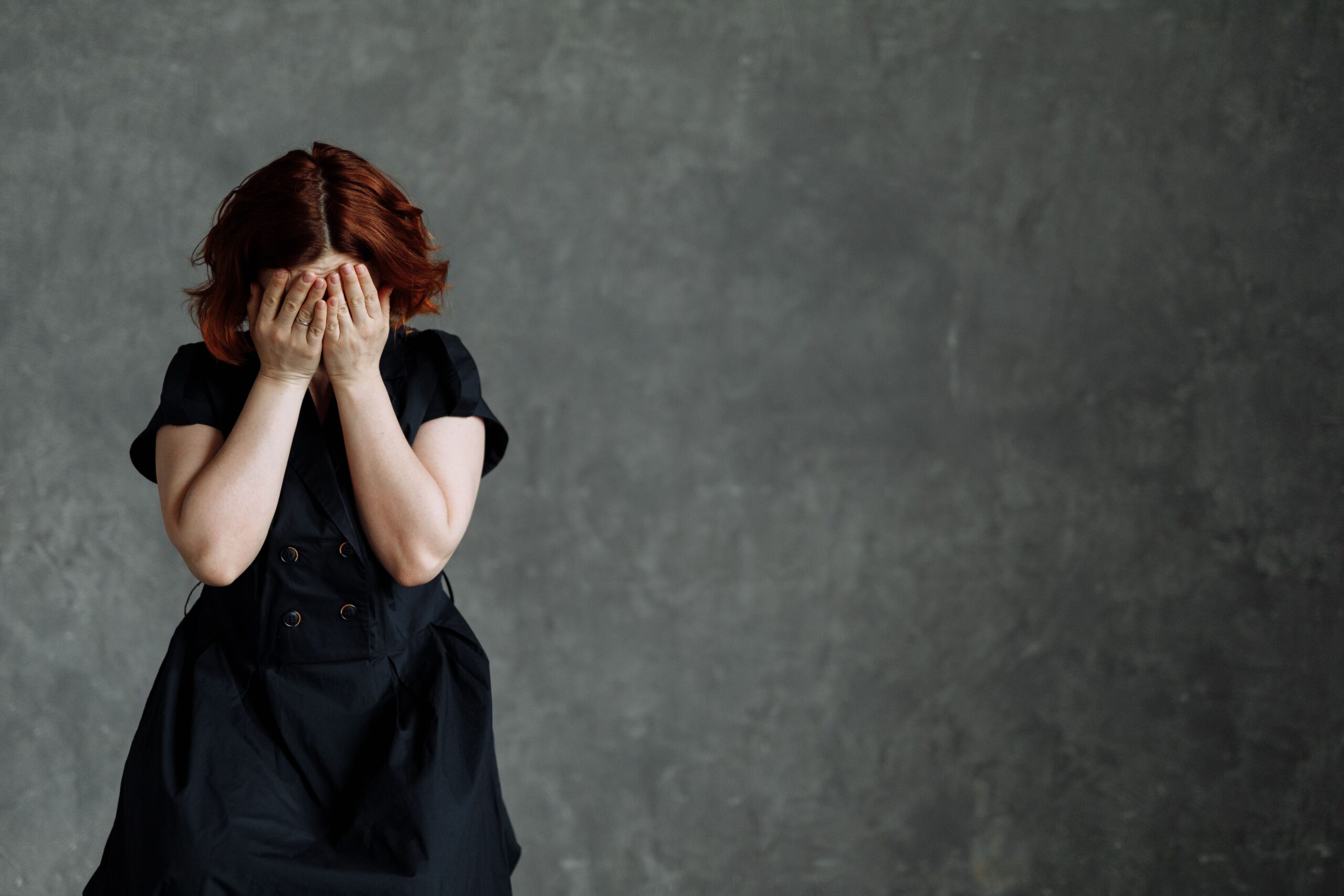 Woman pressing both hands over her face, head bowed, illustrating the physical sensation of a shame response — the body closing in before any thought has caught up.
