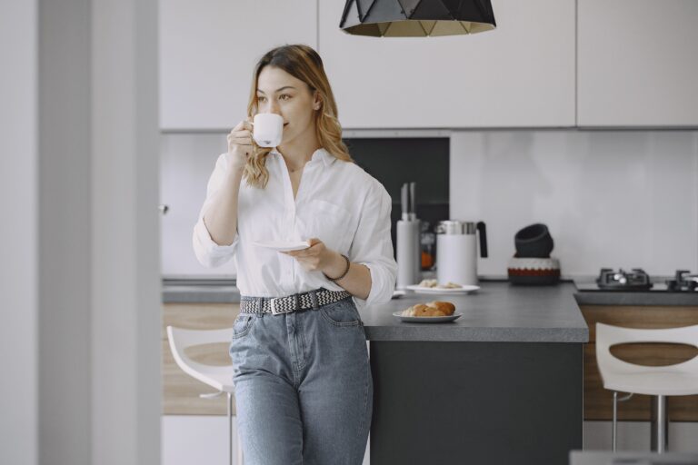 Woman standing in her kitchen drinking coffee in a calm, unhurried moment — illustrating the practice of being ordinary without needing to perform or achieve anything.