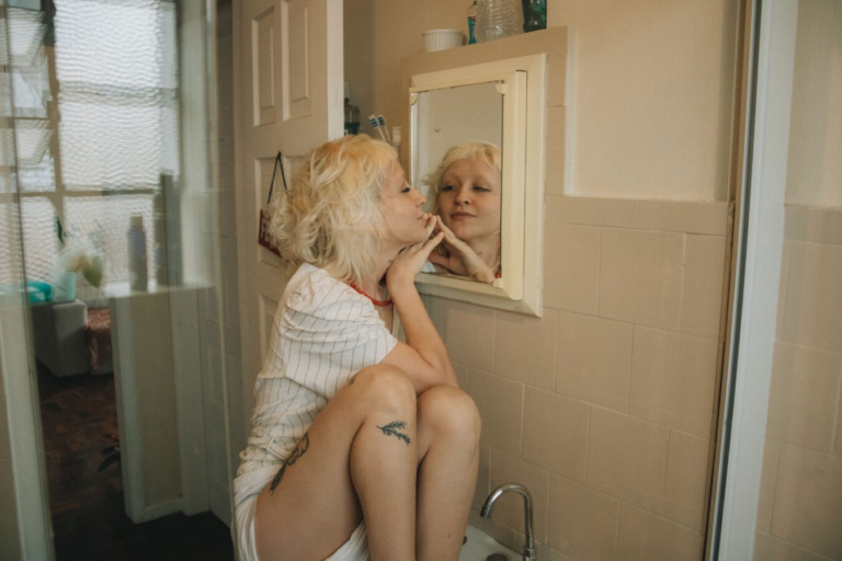 Woman with blonde curly hair sitting on a bathroom sink, resting her chin on her hand and looking closely at her reflection in a small wall mirror.