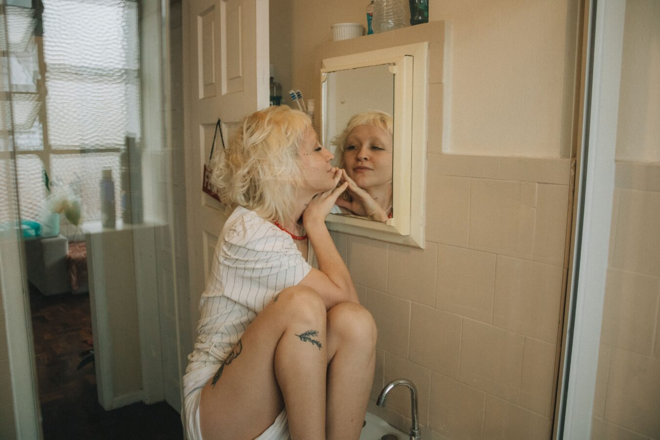 Woman with blonde curly hair sitting on a bathroom sink, resting her chin on her hand and looking closely at her reflection in a small wall mirror.