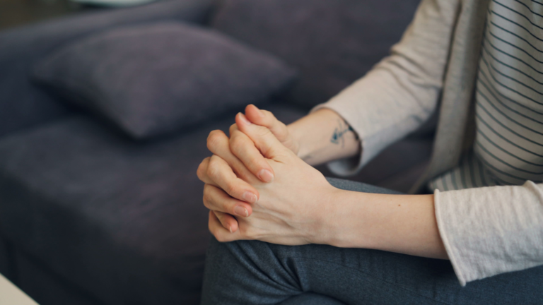Close-up of hands clasped tightly together in someone's lap while sitting on a couch, illustrating the persistent physical tension of background anxiety that stays even in quiet moments.