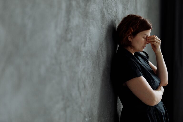 Woman leaning against a wall with eyes closed and hand pressed to her face, illustrating what it feels like to hold in a strong emotion — present on the outside, overwhelmed on the inside.